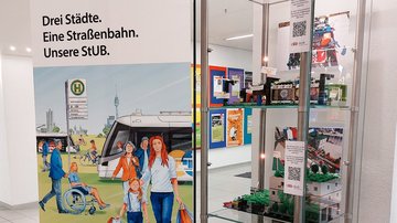 Modell der 6. Lego-Planungswerkstatt in einer Glasvitrine, daneben ein Roll Up zur Stadt-Umland-Bahn.